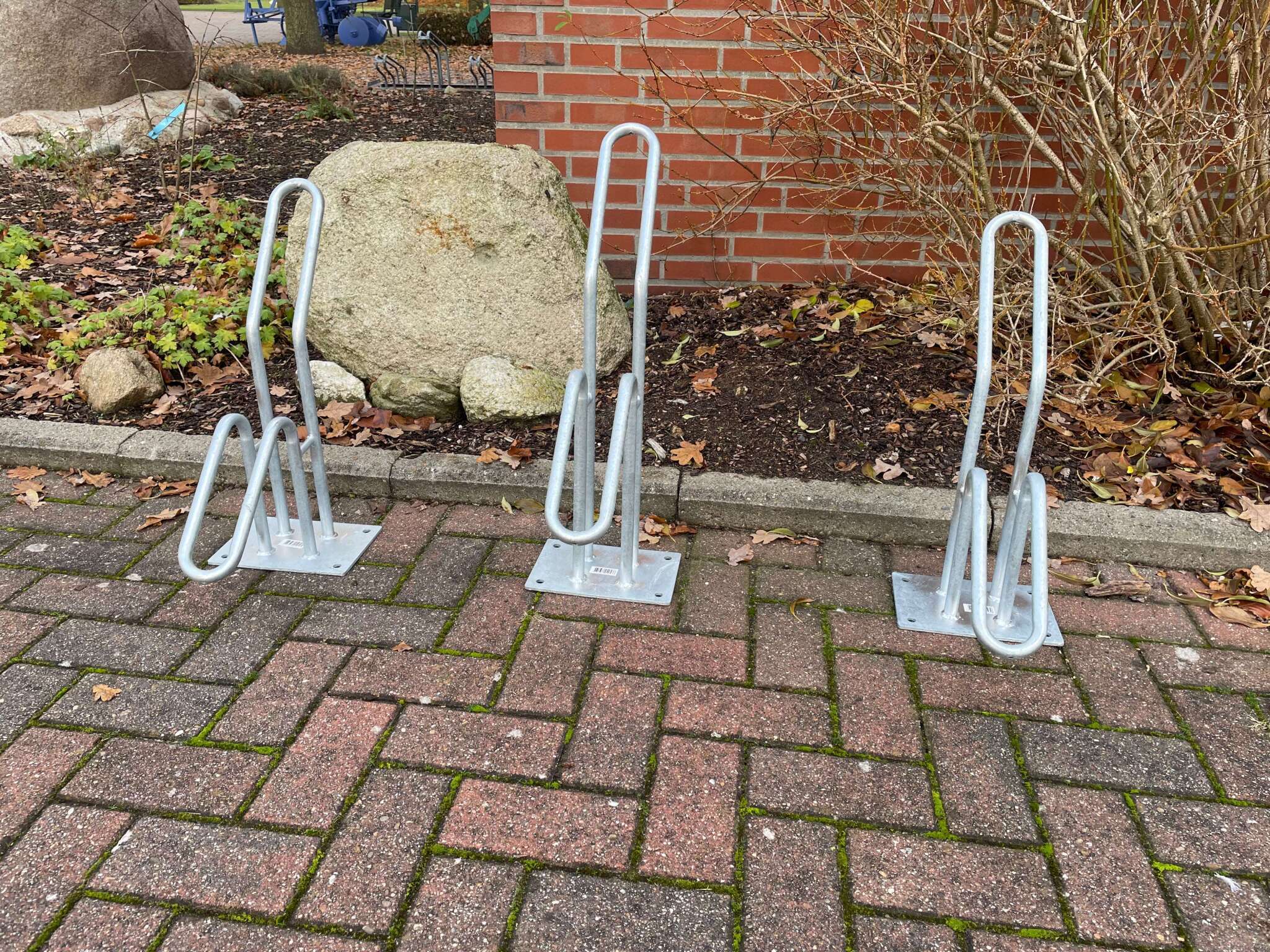 Drei silberne Fahrradständer aus Metall auf einer gepflasterten Fläche vor einem Garten mit Steinen und Laub.