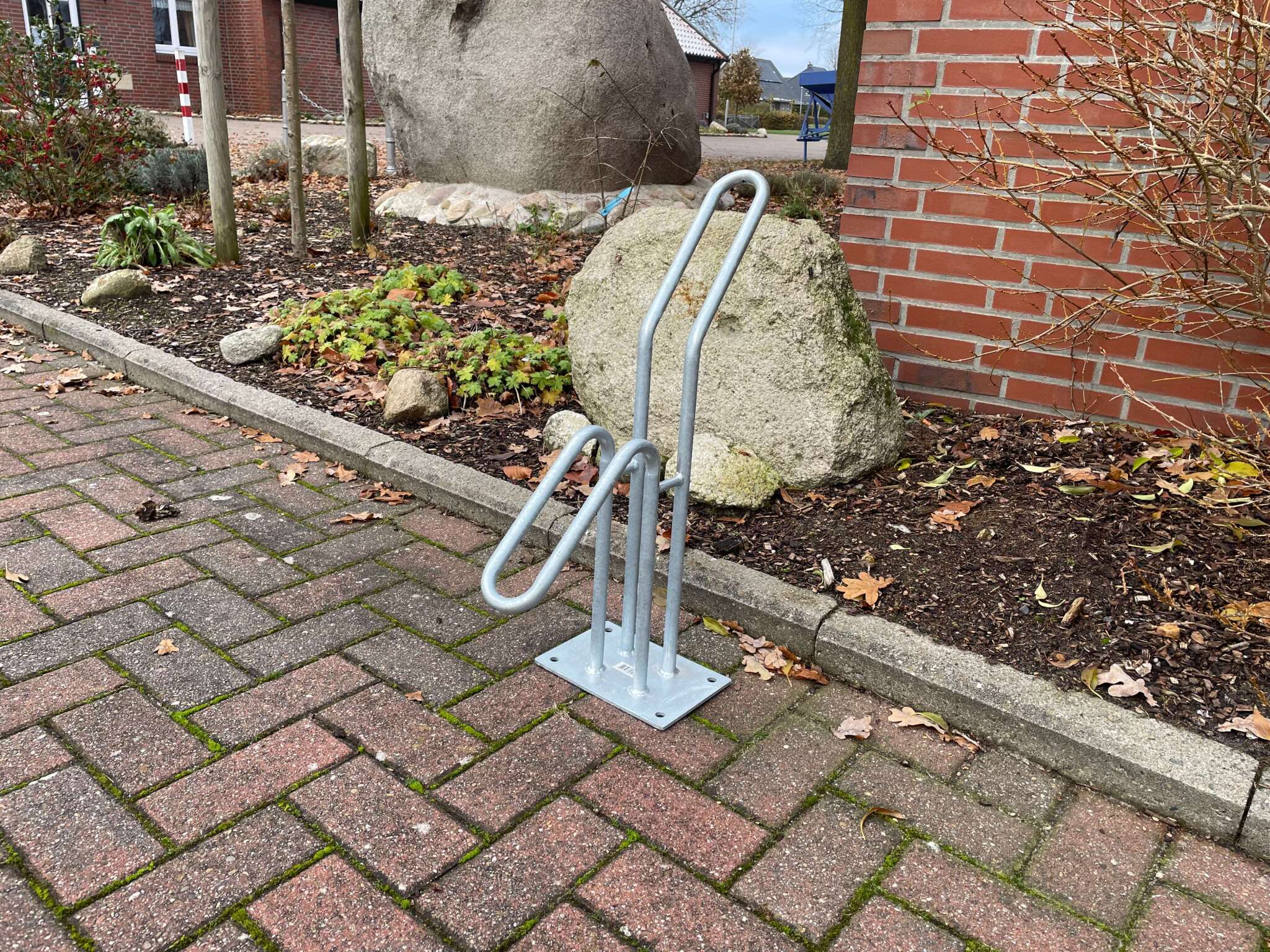 Fahrradständer aus Metall auf gepflastertem Gehweg neben einem Beet mit Steinen und Pflanzen.