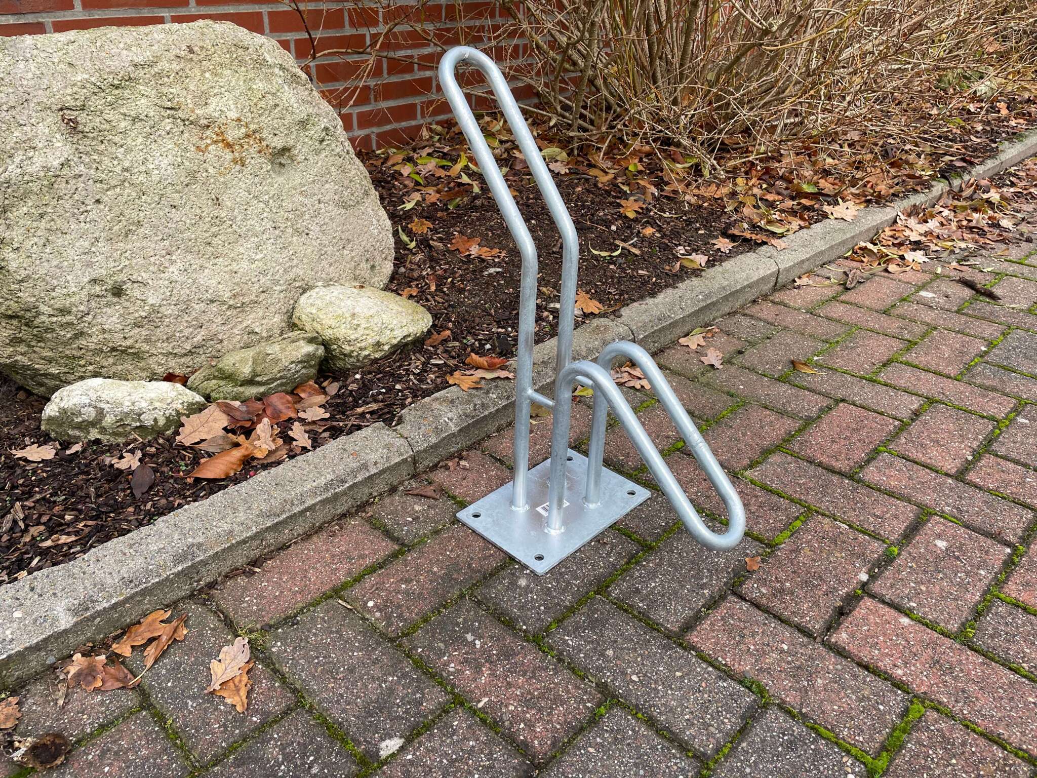 Silberner Fahrradständer aus Metall auf gepflastertem Boden, neben einem Beet mit Steinen und Pflanzen.