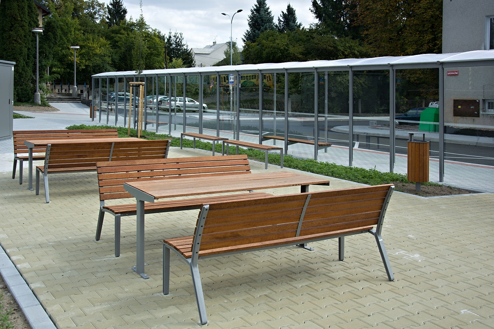 Moderner Außenbereich mit Holzbänken und Tischen auf gepflastertem Platz, umgeben von urbaner Vegetation und Glasüberdachung.