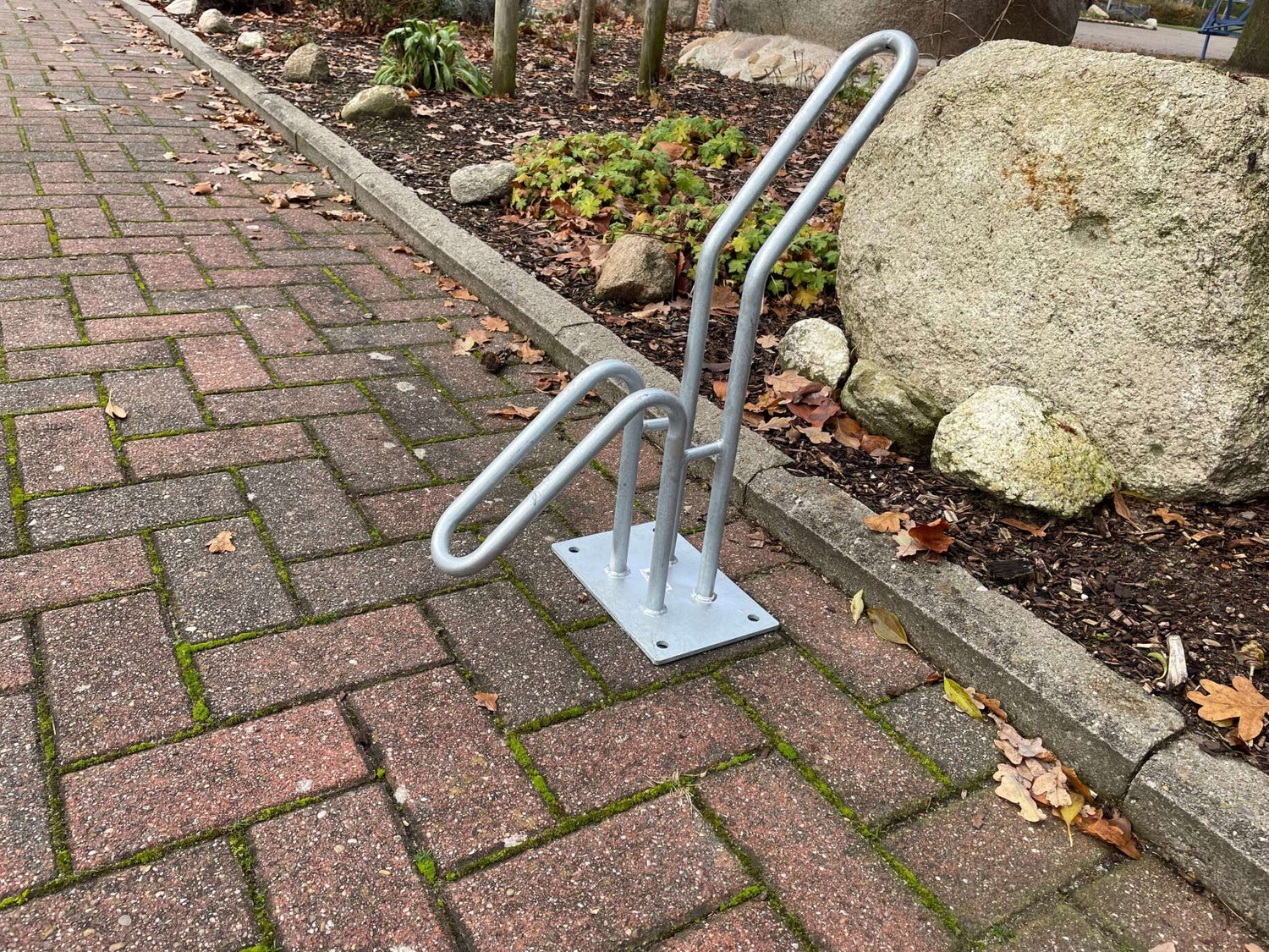 Silberner LIAS Fahrradständer auf gepflastertem Gehweg vor einem Garten mit Felsen und herbstlichen Blättern.