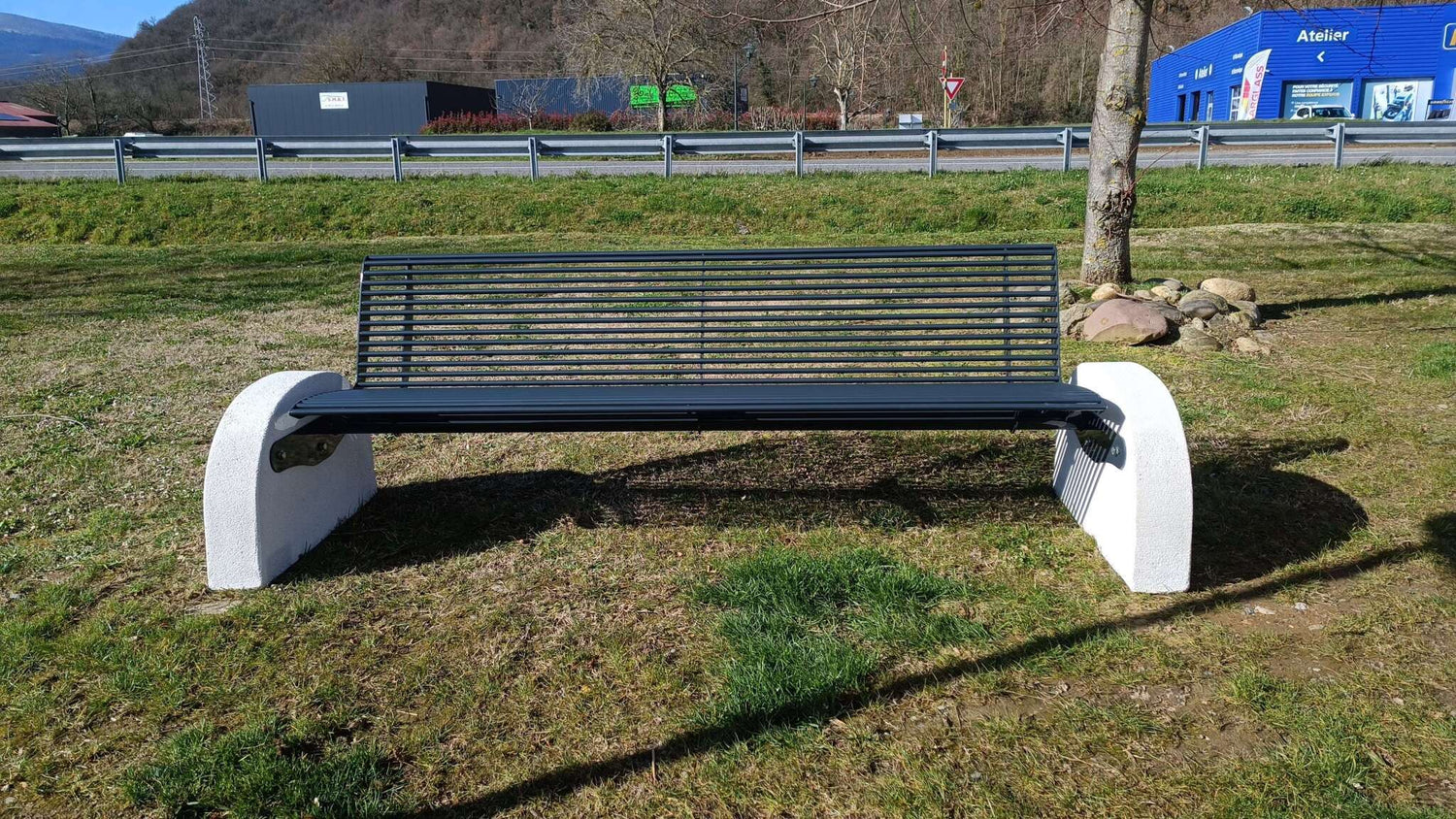 Bank im Freien auf Rasen vor Straße und blauen Gebäuden im Hintergrund.